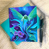 Greeting card featuring artwork by Primal Painter of an ethereal fairy amidst swirling art nouveau flowers and foliage in greens, purples, and blue. 